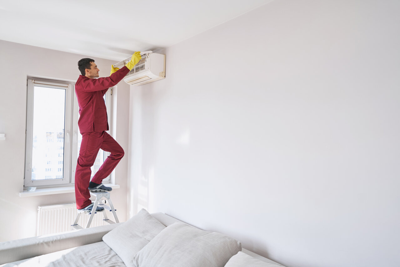 A man in red and yellow attire stands on a ladder, cleaning an air conditioner with focused determination.