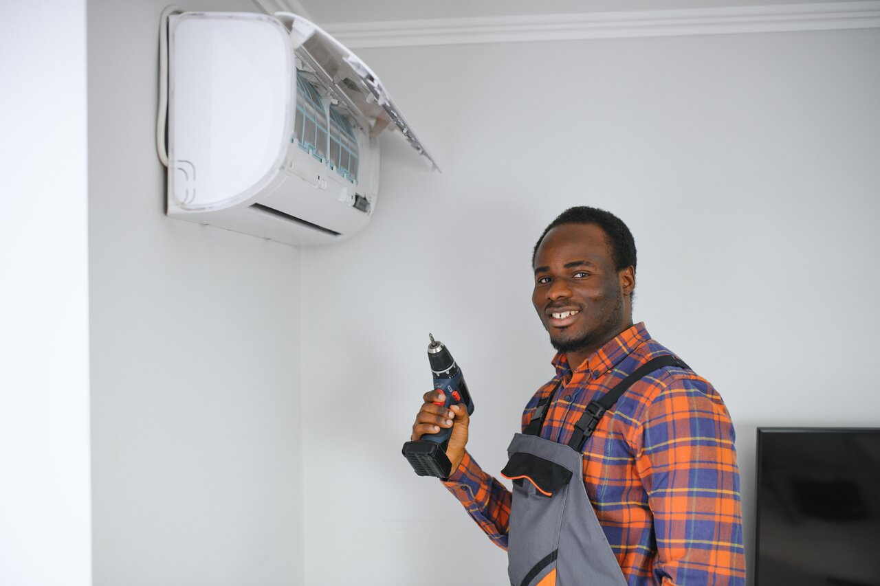 A man operates a drill while holding an air conditioner, demonstrating a home improvement task.