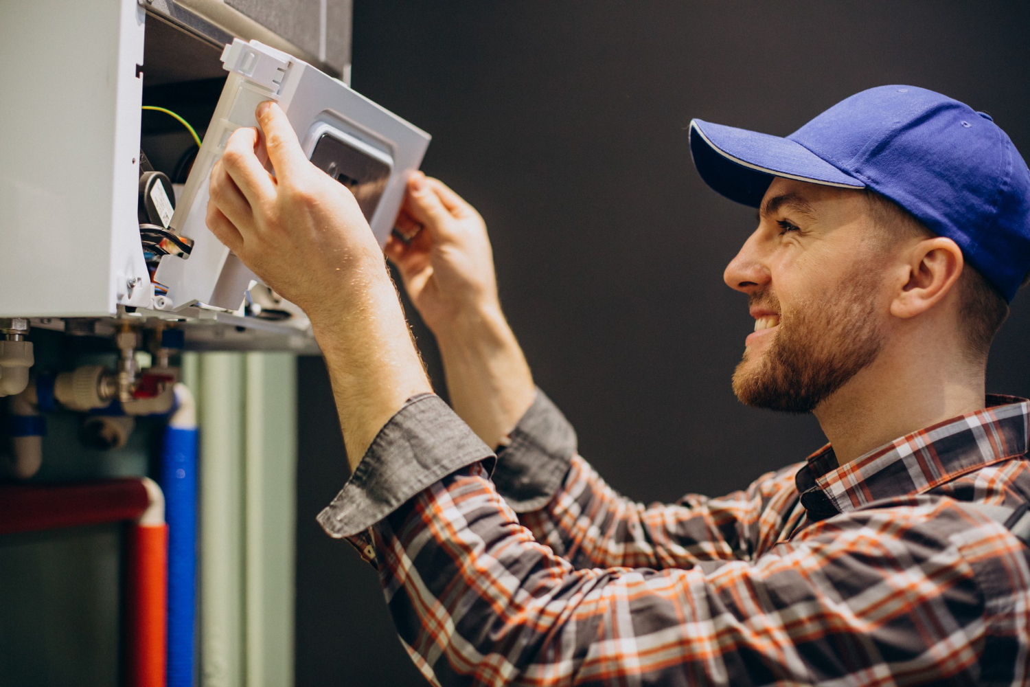 A man wearing a hat and plaid shirt repairs a water heater, demonstrating his skills in home maintenance.