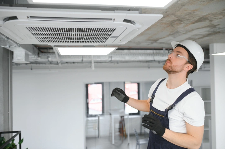 A man wearing overalls and a hard hat stands beside a ceiling fan, showcasing a work environment focused on maintenance.